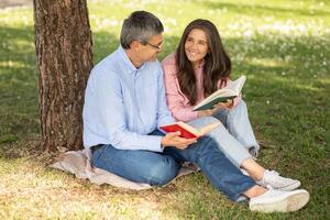 A man and a woman sit together on a blanket, each reading a book under a large tree in a park. They share smiles and laughter while enjoying the warm sunshine. photo