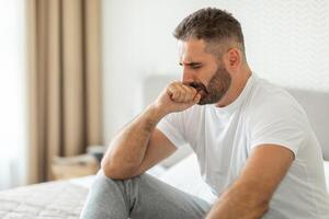A bearded man sits on a bed in a home setting, looking down with a concerned expression, one hand resting on his chin. He wears a white t-shirt and gray pants. photo