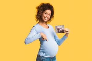 Happy afro pregnant woman with ultrasound scan pointing at tummy over white background photo