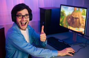A young man wearing headphones sits in front of a computer, playing a game. He is smiling and giving a thumbs-up gesture, indicating his enjoyment of the game. photo