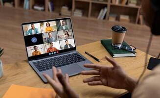 A person is sitting at a desk in front of a laptop computer, participating in a conference call. The laptop screen displays nine people in separate squares. photo