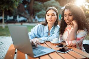 Two young multiethnic women sit at a table outside a cafe, working on a laptop. The image captures a common scene of people working remotely or collaborating on a project. photo