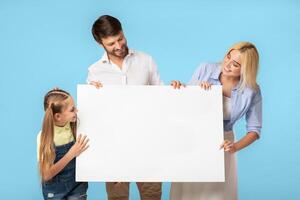 A family consisting of a mother, father, and daughter, are standing in front of a blue background. They are all smiling and holding a large white poster in front of them. photo