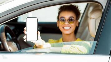 African American woman is sitting in the drivers seat of a car with the window down. She is smiling and holding a smartphone with a blank screen towards the camera. photo