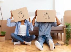 Unrecognizable Afro Couple Wearing Moving Boxes With Smiley Faces On Head Having Fun Sitting On Floor In New Home photo