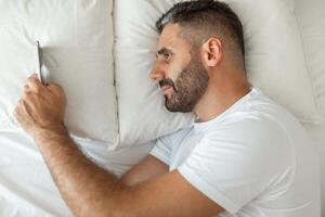 A man lies in bed, propped up on one elbow, his head resting on a pillow. He is looking at a smartphone with his right hand while his left arm is resting on the white bed sheet. photo