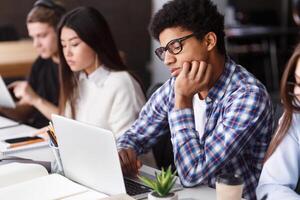 A multiethnic group of focused young adults is sitting at desks in a classroom setting. They appear to be absorbed in their individual tasks, with some using laptops and others taking notes photo