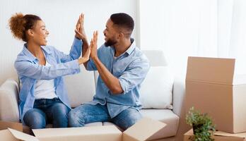 House Owners. Happy Afro Spouses Giving High-Five Sitting Among Unpacked Moving Boxes In Own Apartment. Copy Space photo