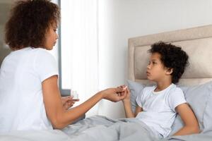 A concerned African American mother sits on the edge of a bed, gently holding her young child hand as she checks for a fever. The child, looking unwell photo