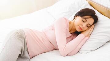 A woman is reclining on a bed, resting her head on a soft pillow. She appears relaxed and comfortable in her surroundings. The room is simple and tidy, with minimal decor. photo
