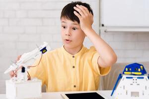 A young boy sits at a table, looking at his robot arm project with a frustrated expression. He has one hand on his forehead, and the other is holding the robot arm, which appears to be malfunctioning photo