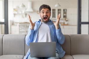 A man with a shocked expression sits on a couch in his home with a laptop in his lap. He is surprised and excited by something on the screen. photo