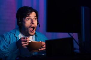 A young man wearing a headset and a blue shirt sits in front of a computer screen, looking at the screen with a shocked expression on his face while holding a bowl of cereal and a spoon in his hands photo
