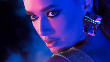 A woman stares intensely at the camera under dramatic blue and purple studio lighting. She wears bold makeup and a large metallic earring. The setting suggests a nighttime atmosphere photo
