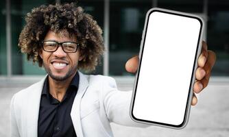 Black man standing and holding a smartphone in front of him, displaying a blank white screen. The mans hand is clearly visible as he holds the device upright. photo