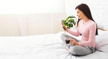 A young woman sits cross-legged on a white bed, holding a coffee mug in one hand and a tablet in the other. She seems engrossed in her digital device, perhaps reading or browsing, copy space photo