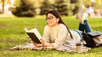 Asian woman is lying on the grass, engrossed in reading a book. She is surrounded by nature, with trees and flowers in the background. The sun is shining, casting a warm glow over the scene. photo