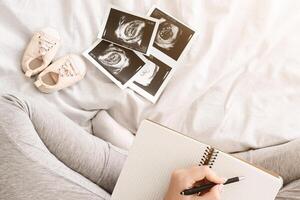An expectant mother is sitting on a bed writing in a journal. Surrounding her are baby ultrasound images and a pair of tiny baby shoes, capturing a quiet moment of anticipation and planning photo