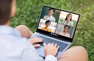 A woman sits on the grass in a park, using a laptop to participate in a conference call with four other people, over shoulder view photo