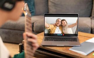 A young guy is seen playing an acoustic guitar while chatting with an older couple on a laptop. The couple is smiling and waving, seemingly enjoying the music. photo