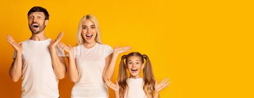 A joyful family stands in front of a vibrant yellow backdrop, with each member expressing excitement and happiness through wide smiles and raised hands, copy space photo
