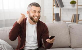 A man is seated on a couch, his hand gripping a remote control. The man appears relaxed and focused on the TV. The room is cozy, with soft lighting enhancing the scene. photo