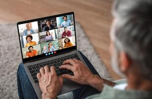 Mature man sits on a couch at home, using a laptop to participate in a virtual meeting. He is focused on the screen, which displays a grid of feeds of other participants photo
