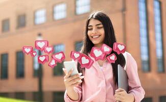 A young woman with long, dark hair is smiling while looking at her smartphone. She is standing in front of a university building, and several heart icons are floating around her phone photo