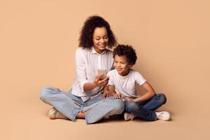 African American woman and a child are seated on the floor, both looking intently at phone screen. The woman appears engaged in conversation or viewing content, while the child watches attentively. photo