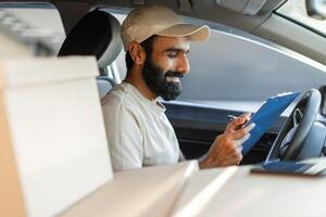 Indian courier checks his delivery list while sitting in the driver seat of his vehicle. Several packages are visible inside the vehicle, indicating a busy delivery schedule. photo
