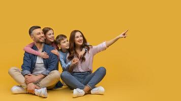 A family consisting of parents and two children is sitting on the floor indoors. They are all pointing at an object or an event happening in front of them. photo