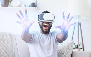 African American man wearing a virtual reality headset and interacting with the virtual world. He is sitting on a couch and has his hands extended outwards as if he is reaching for something photo