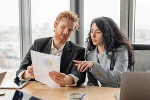 A man and a woman in business attire are reviewing a data graph during a meeting in a modern office. The man is seated at a table, holding the graph and looking at it intently photo