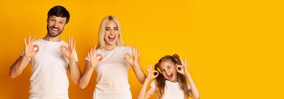 A cheerful family consisting of a father, mother, and daughter stands together, all smiling widely while making the okay hand gesture. Their playful expressions and matching casual outfits photo