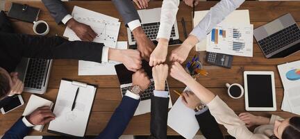 Young people putting their fists together as symbol of unity and achievement, top view. Group of people fist bump assemble together over workplace. Teamwork concept, copy space in middle photo