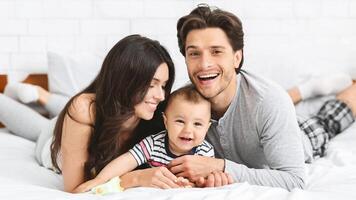 This image shows a happy family of three, a man, woman, and baby, lying on a white bed. The parents are smiling, and the baby is laughing. They are all wearing casual clothing photo