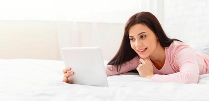 A woman is lying comfortably on a white bed, smiling as she uses a tablet device. The cozy bedroom features soft natural light coming through the window, creating a peaceful and inviting atmosphere. photo