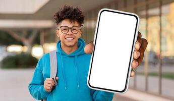 A young black guy with curly hair and glasses is smiling at the camera while holding up a smartphone with a blank screen, standing in front of a university building photo