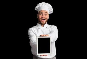 Excited Chef Showing Digital Tablet With Empty Screen Recommending Restaurant Menu Or Food Delivery Service Posing On Black Studio Background. photo