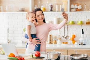 A woman is holding a baby in her arms while taking a selfie with her smartphone. Both mother and child are looking at the camera, smiling. The background is blurred. photo