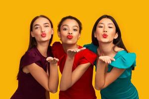 Three Beautiful Multiethnic Girls Sending Air Kiss Looking At Camera Having Fun Standing In Studio Over Yellow Background. Panorama photo