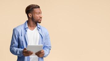 Serious Afro Guy Holding Digital Tablet Looking Aside Standing Over Yellow Studio Background. Panorama, Copy Space photo