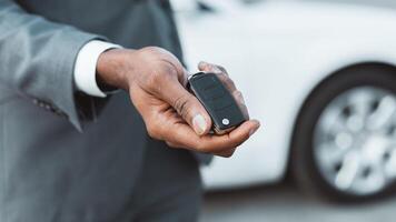 A man in a grey suit is holding a car key in his right hand. He is standing in front of a white car, and the key is in focus while the car is out of focus in the background, copy space photo