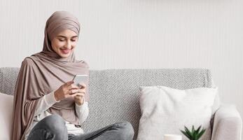 Smiling muslim girl in headscarf using smartphone at home, messaging or browsing social networks, relaxing on couch in living room, copy space photo
