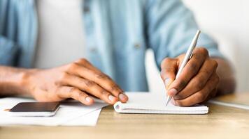 A person sits at a desk writing in a notebook with a pen. Their left hand is resting on a phone on the table. photo