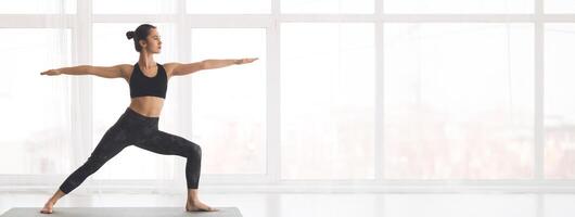 A person performs the warrior pose in a well-lit studio, showcasing strength and balance against a backdrop of large windows, copy space photo