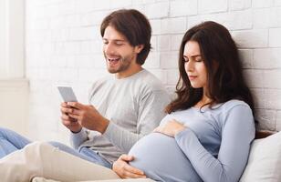 Indifferent husband. Future father using smartphone ignoring his pregnant wife that suffering belly ache on bed at home, free space photo