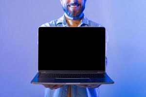 A smiling man with a beard stands against a blue background, holding a laptop with the blank screen facing forward. He appears cheerful and enthusiastic, cropped photo