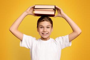 Positive schoolboy holding stack of books on his head, orange studio background photo
