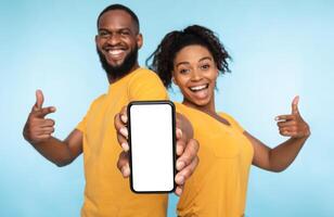 Use this new application. Cheerful black couple pointing at smartphone with blank screen, recommending mobile app, advertising your website, mockup for design. Selective focus photo